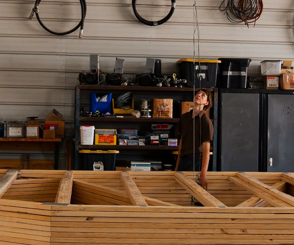 A man in a workshop, standing next to a stack of trusses and looking up